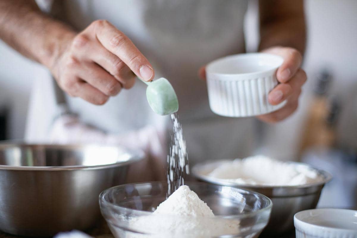 Seseorang menambahkan bahan kering ke dalam mangkuk kaca berisi tepung menggunakan sendok takar di dapur.