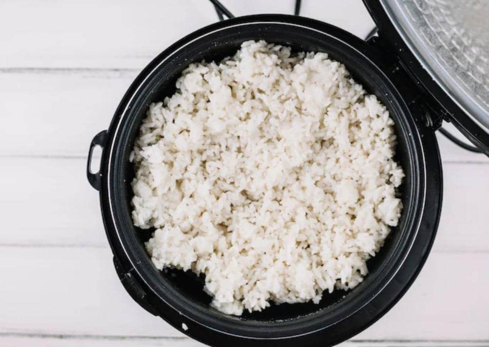 Nasi putih yang sudah matang di dalam rice cooker hitam dengan tutup terbuka di atas meja berwarna terang.