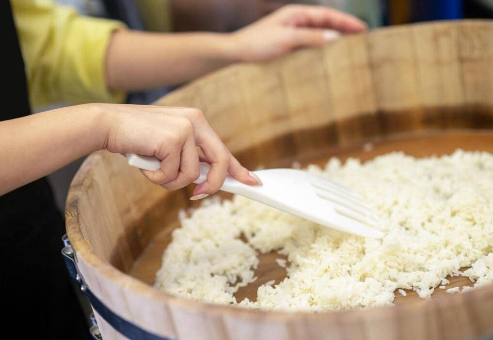 Tangan seseorang mengaduk nasi putih menggunakan spatula plastik putih di dalam wadah kayu besar.