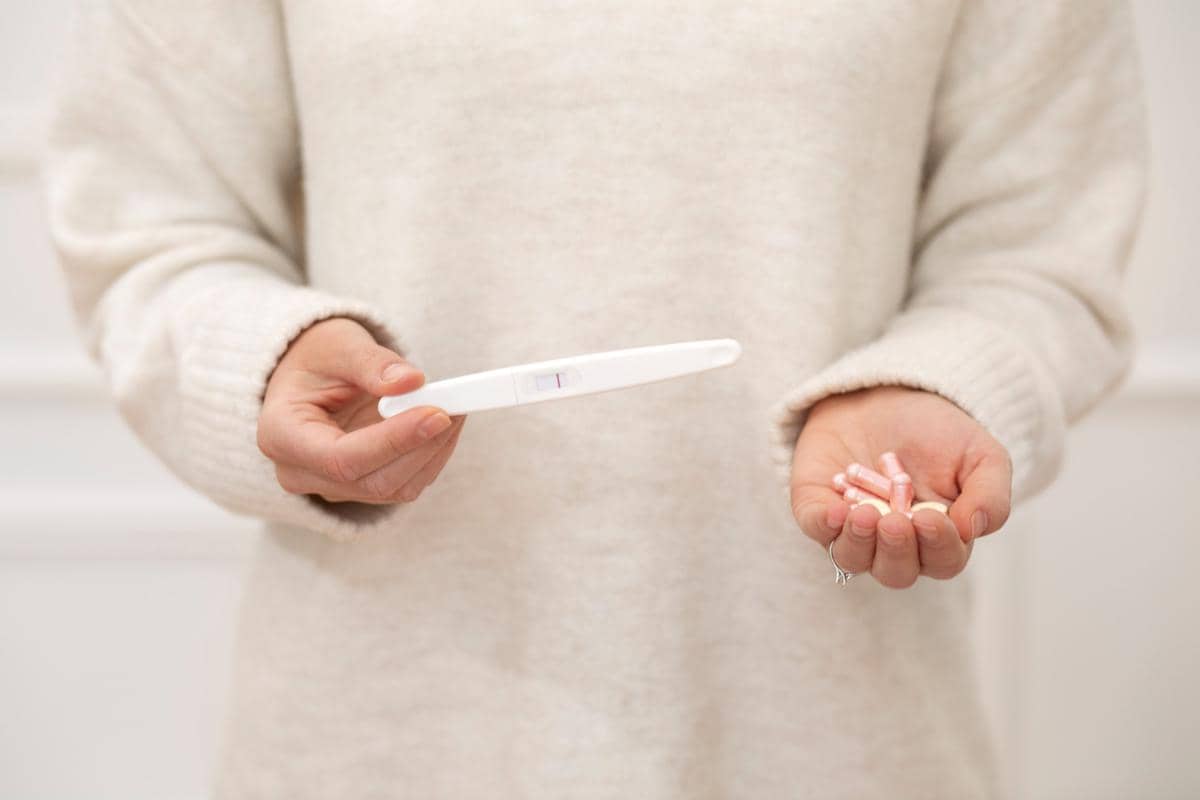 Seseorang mengenakan sweter krem memegang alat tes kehamilan dengan hasil negatif di satu tangan dan beberapa kapsul obat di tangan lainnya.