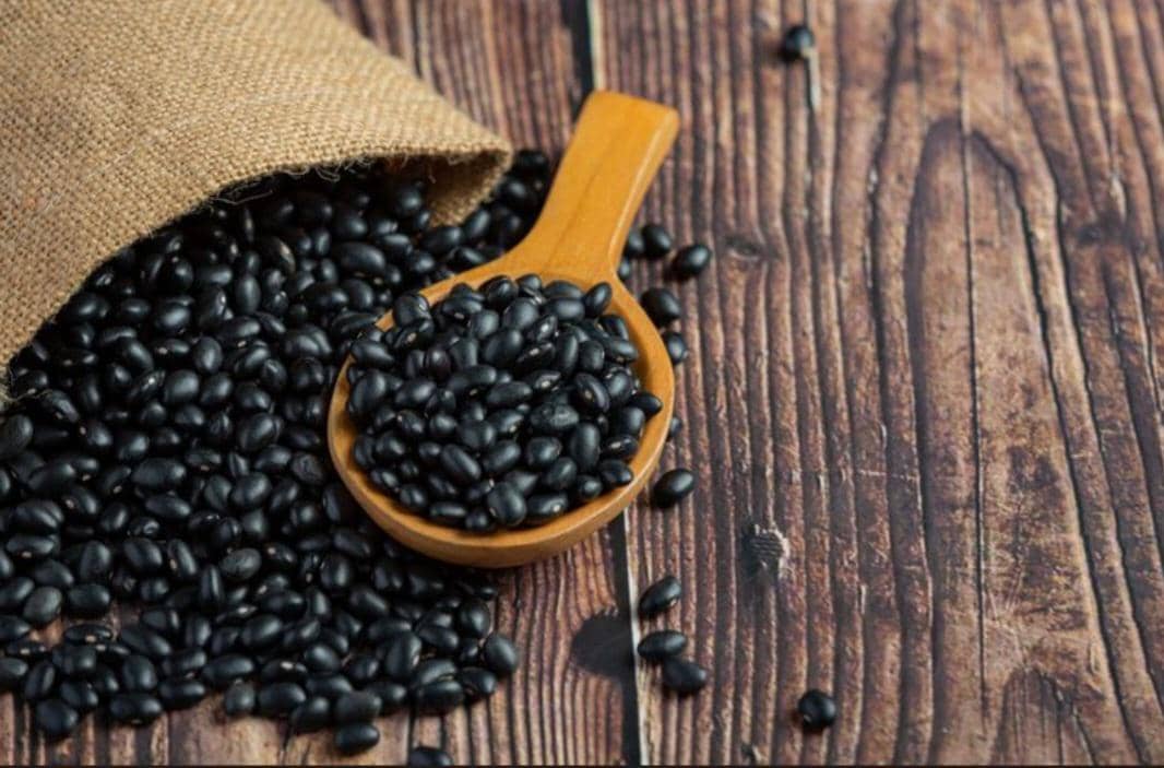 Kedelai hitam kering tumpah dari karung goni kecil dengan sebagian diambil menggunakan sendok kayu di atas meja kayu.