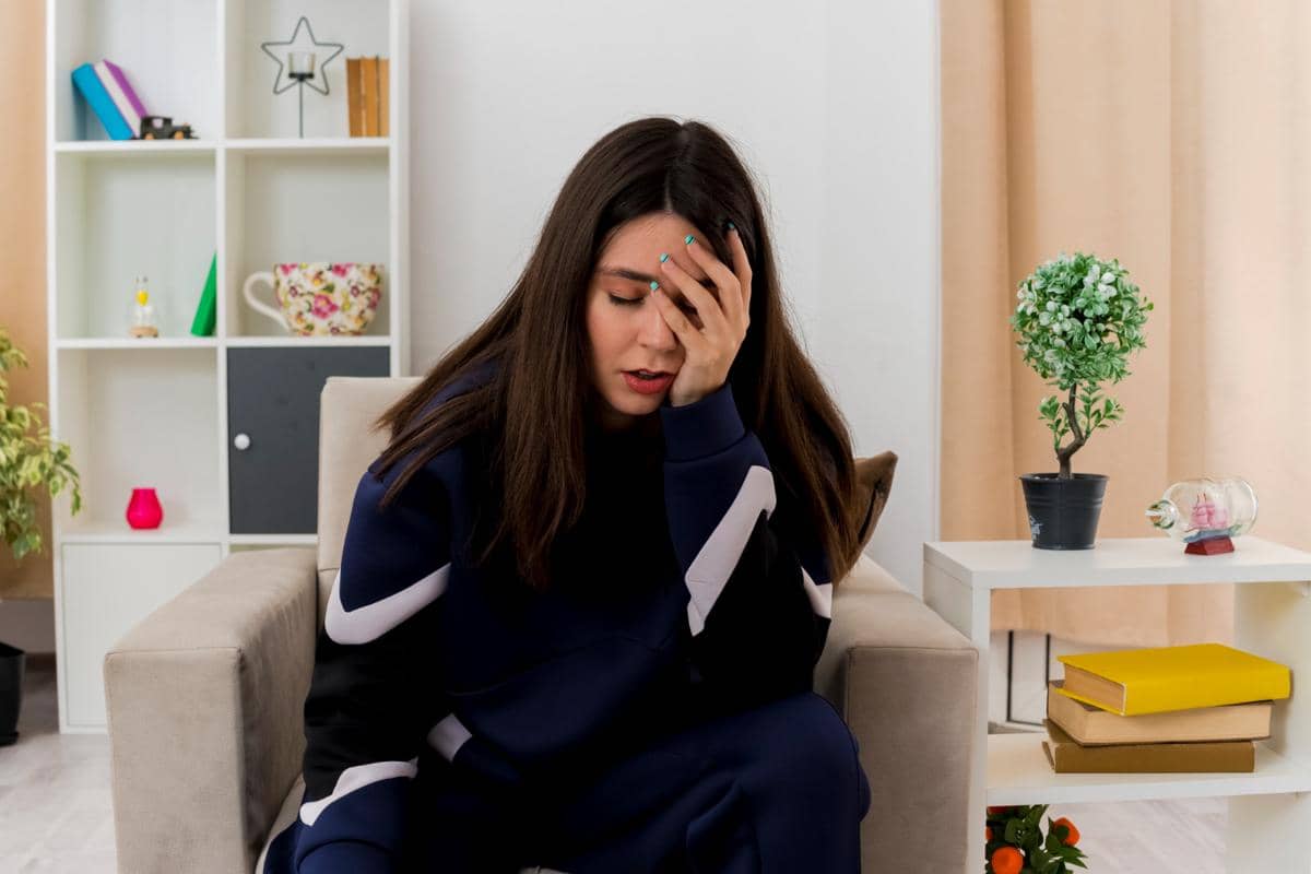 Seorang wanita duduk di ruang tamu dengan tangan menutup wajah, tampak lelah dan tertekan