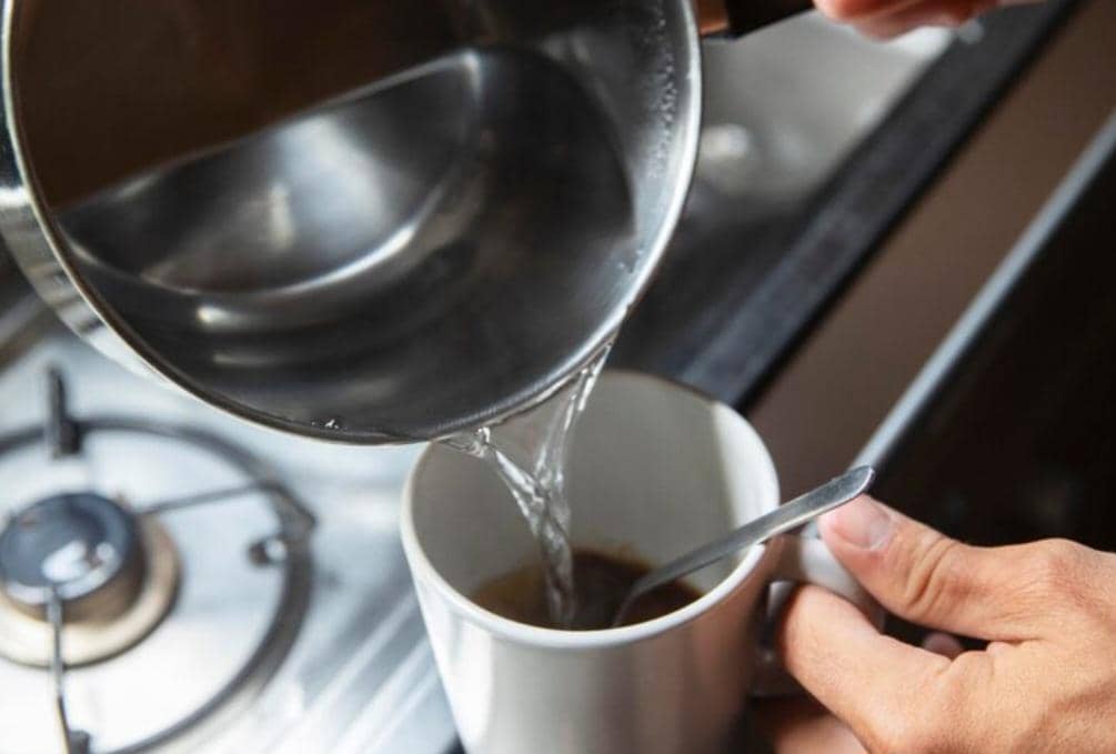 Seseorang menuangkan air panas dari panci ke dalam cangkir berisi bubuk kopi di dapur dengan kompor gas di latar belakang.