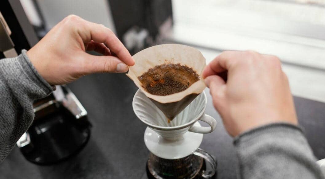 Tangan seseorang memegang kertas saring berisi ampas kopi di atas alat penyeduh kopi manual di meja dapur.