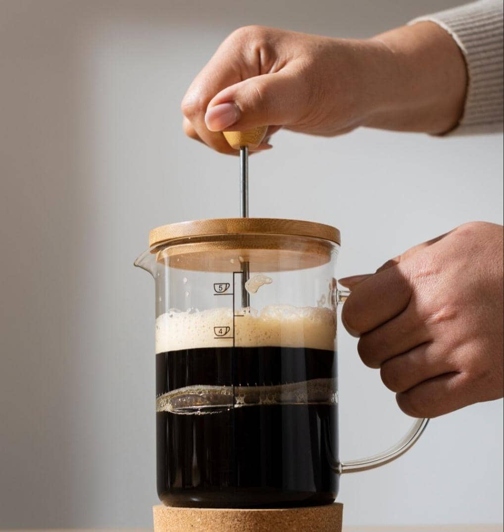 Tangan seseorang menekan tuas pada alat French press kaca berisi kopi hitam dengan busa di bagian atas di atas meja.