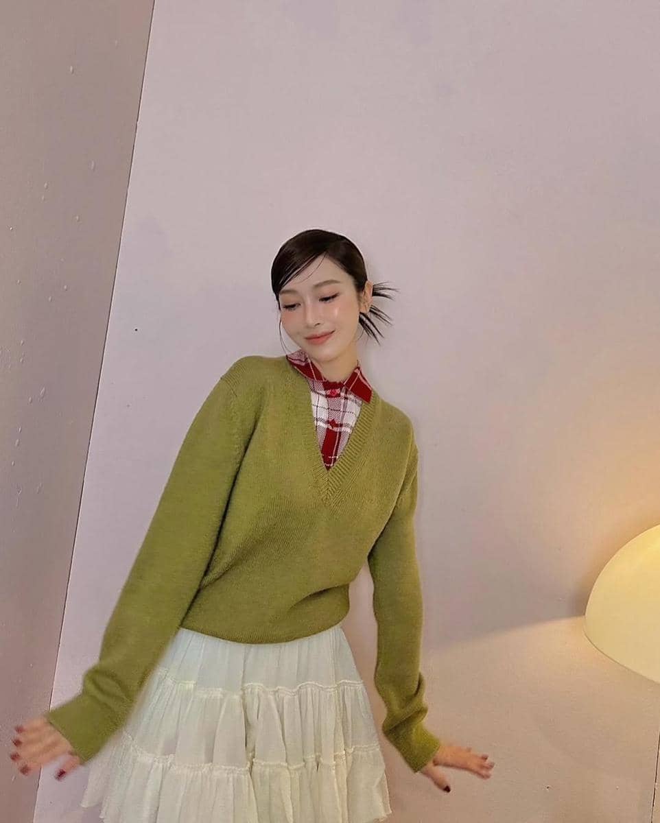 Seorang wanita mengenakan sweater hijau zaitun dengan kemeja kotak-kotak merah dan rok putih berlapis, berpose di dalam ruangan.