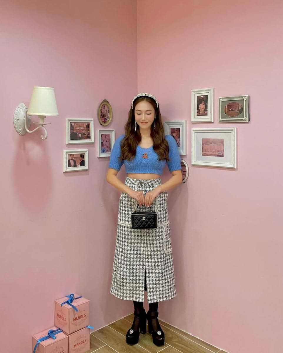 Seorang wanita mengenakan crop top biru dan rok motif houndstooth hitam putih, memegang tas kecil hitam di ruangan bernuansa pink.