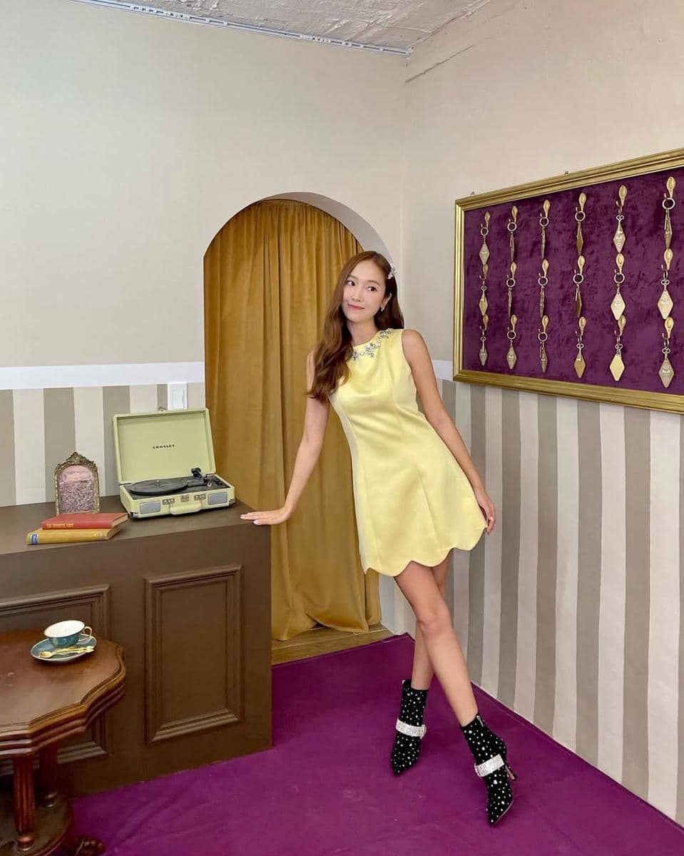 Seorang wanita mengenakan gaun kuning pastel tanpa lengan dan sepatu hitam berhias mutiara berpose di ruangan bergaya retro dengan tirai emas.