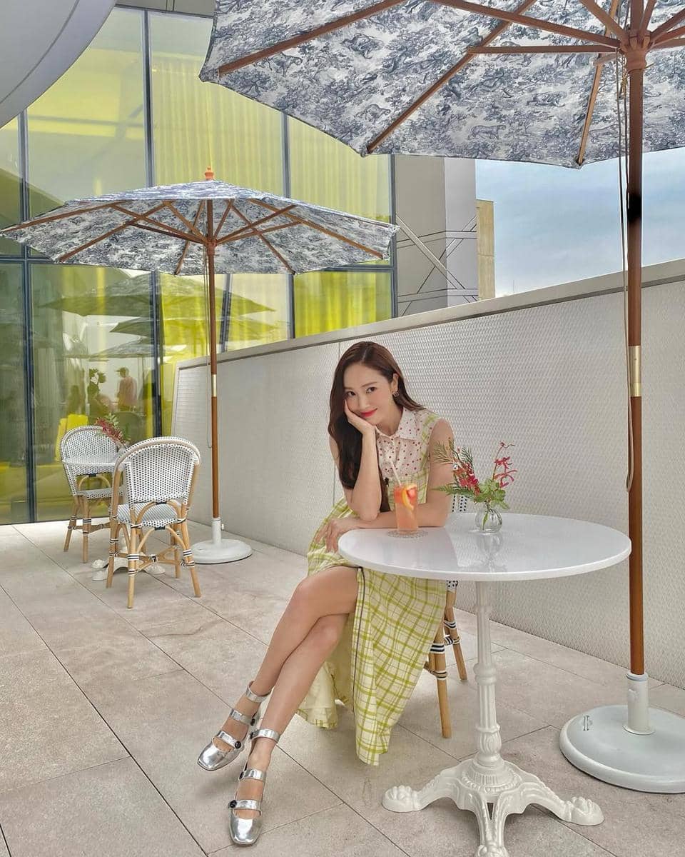 Seorang wanita duduk santai di kafe outdoor dengan meja putih, mengenakan gaun bermotif kotak hijau dan sepatu perak berkilau.
