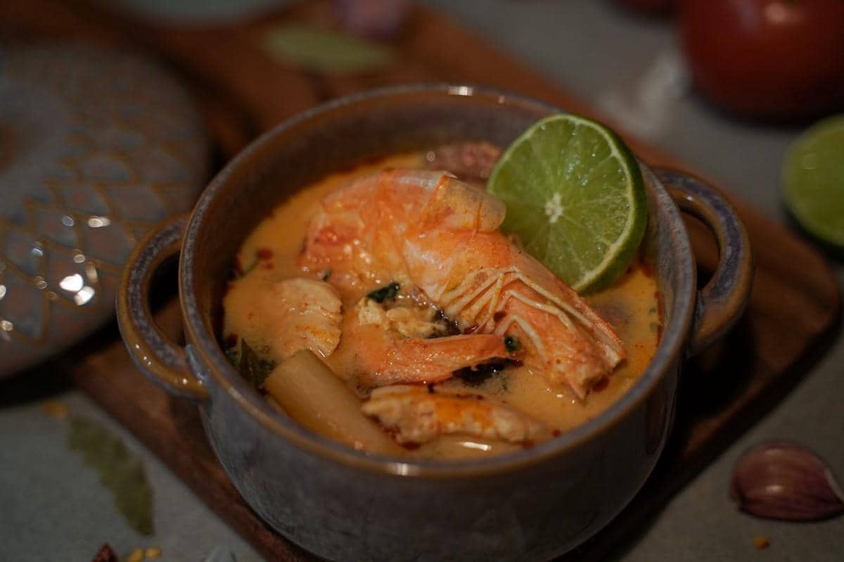 Semangkuk tom yum udang dengan kuah berwarna oranye, potongan jeruk nipis, dan rempah-rempah tersaji dalam mangkuk keramik.