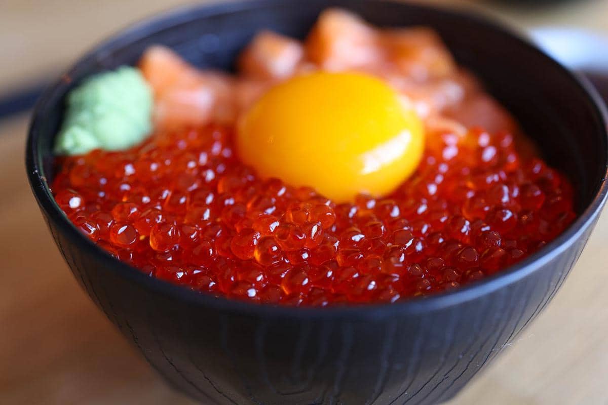 Semangkuk donburi berisi nasi dengan aneka topping, disajikan di atas meja kayu bersama peralatan makan.