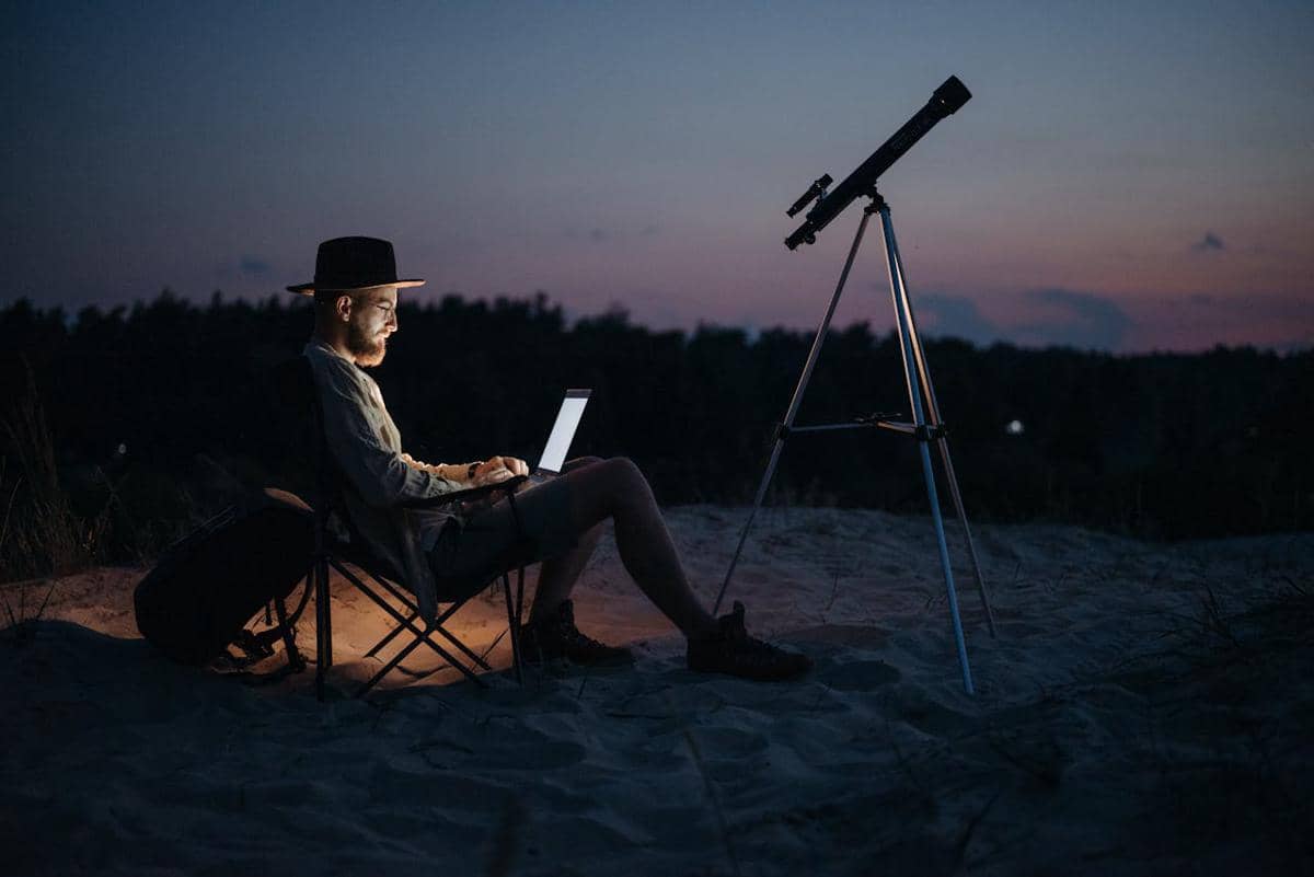 seseorang yang sedang duduk melihat laptop dengan sebuah teleskop di depannya untuk mengamati benda langit
