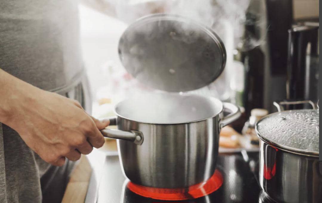 Seseorang memegang panci stainless di atas kompor listrik dengan air mendidih dan uap panas keluar dari dalam panci.