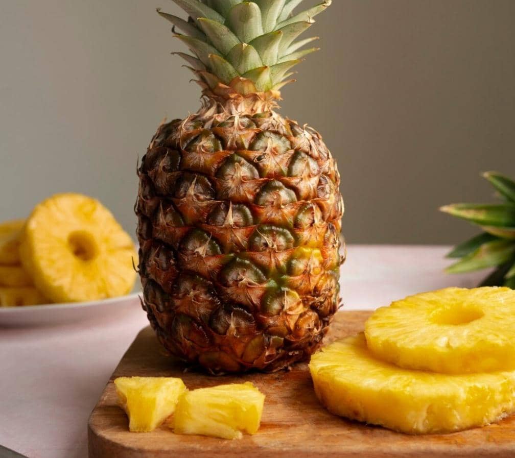 Nanas matang utuh dengan potongan nanas segar di atas talenan kayu, menampilkan tekstur kulit dan daging buah berwarna kuning cerah.