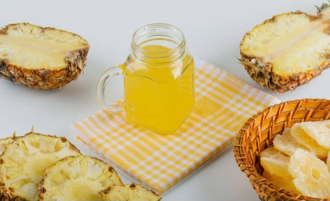 Segelas air perasan nanas berwarna kuning diletakkan di atas kain bermotif kotak kuning putih, dikelilingi potongan nanas segar dan kering.