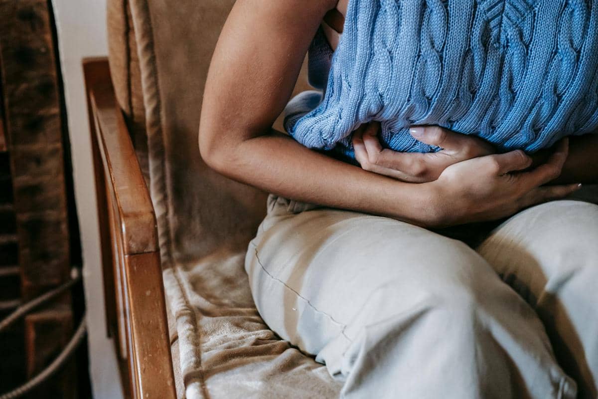 Seseorang duduk di kursi sambil memegangi perut dengan bantal rajut biru, menggambarkan rasa sakit perut atau kram.