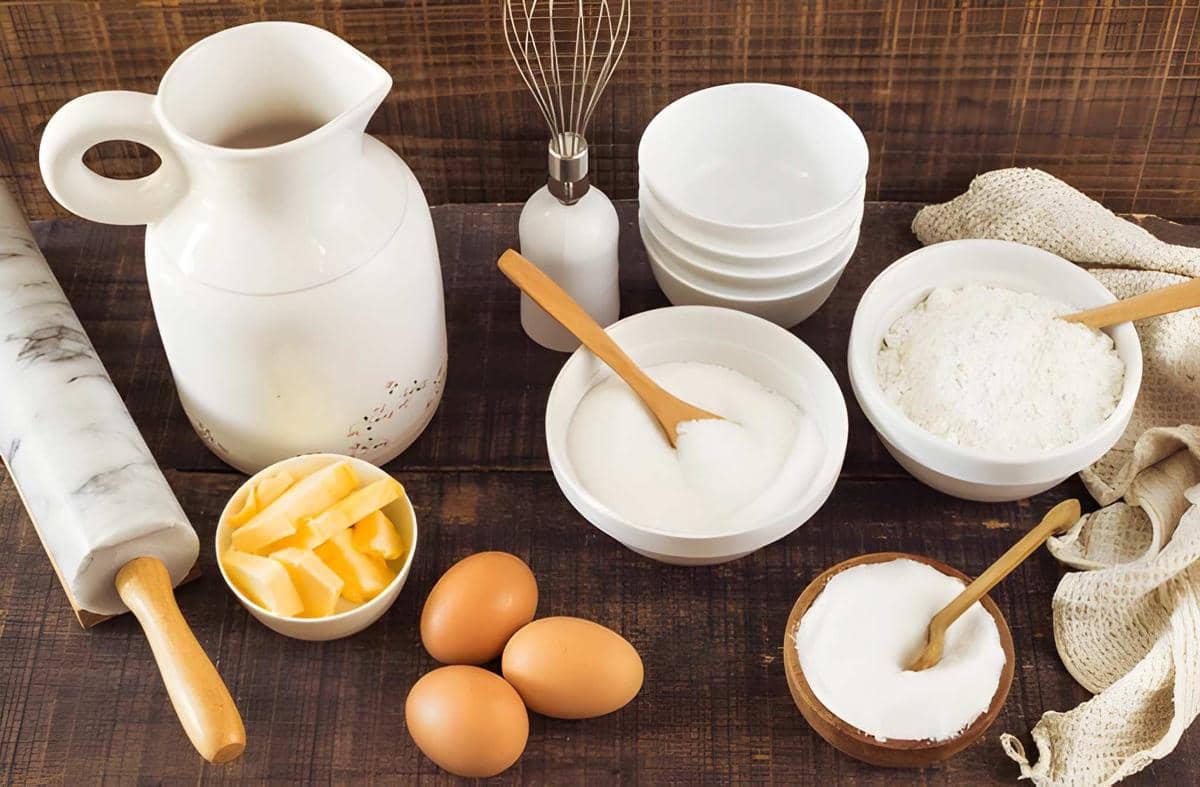 Aneka bahan untuk membuat kue, termasuk tepung, telur, mentega, gula, dan alat pengaduk di meja dapur.