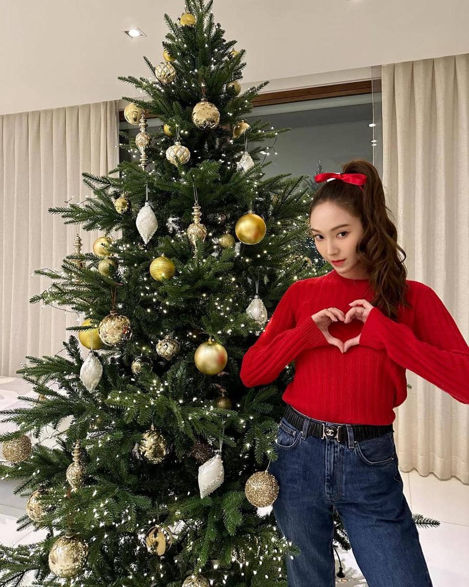 Seorang wanita mengenakan sweater merah dan celana jeans berpose dengan tangan membentuk hati di depan pohon Natal berhias ornamen emas.