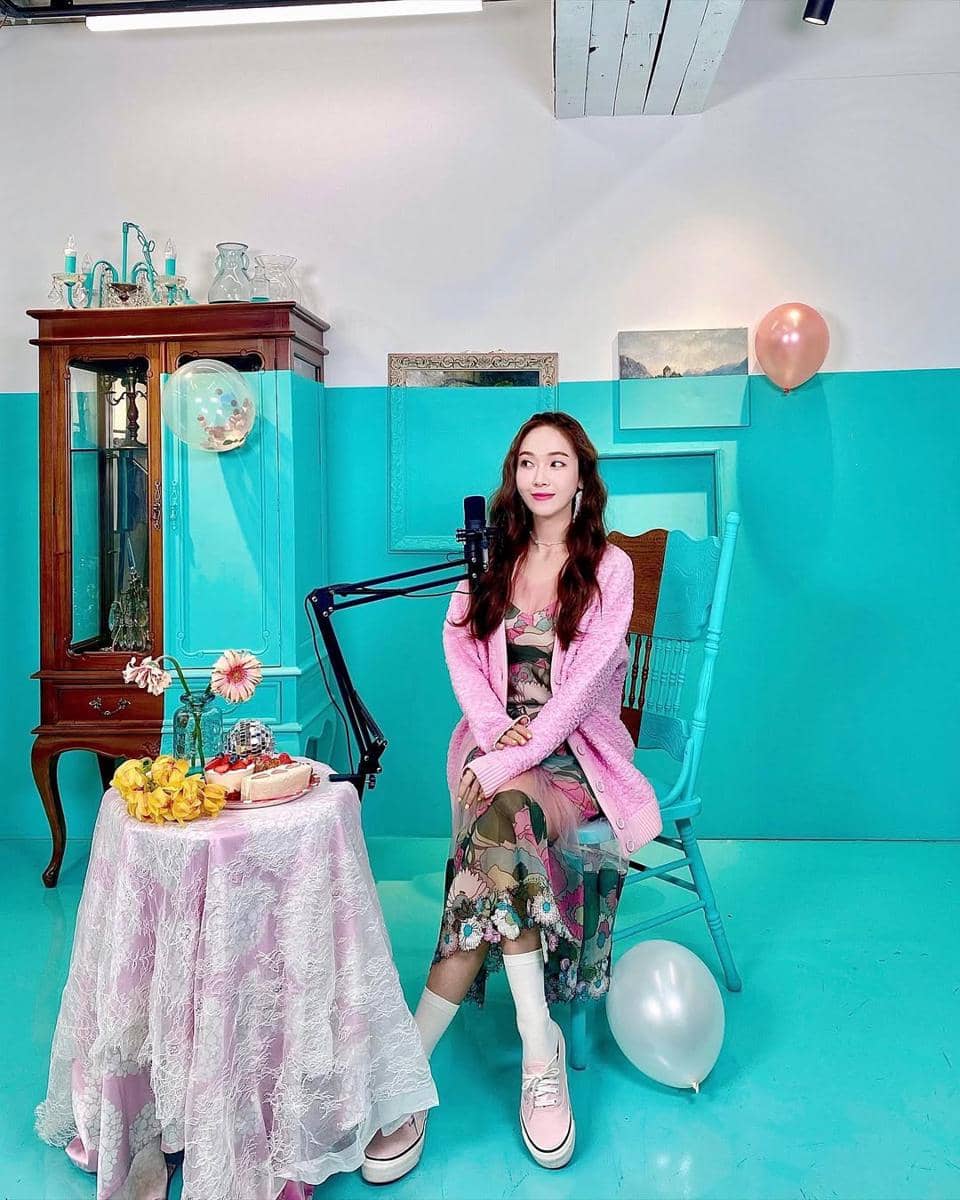 Seorang wanita duduk di kursi biru mengenakan gaun bermotif bunga dan kardigan pink di ruangan bernuansa biru pastel dengan dekorasi balon dan meja bunga.