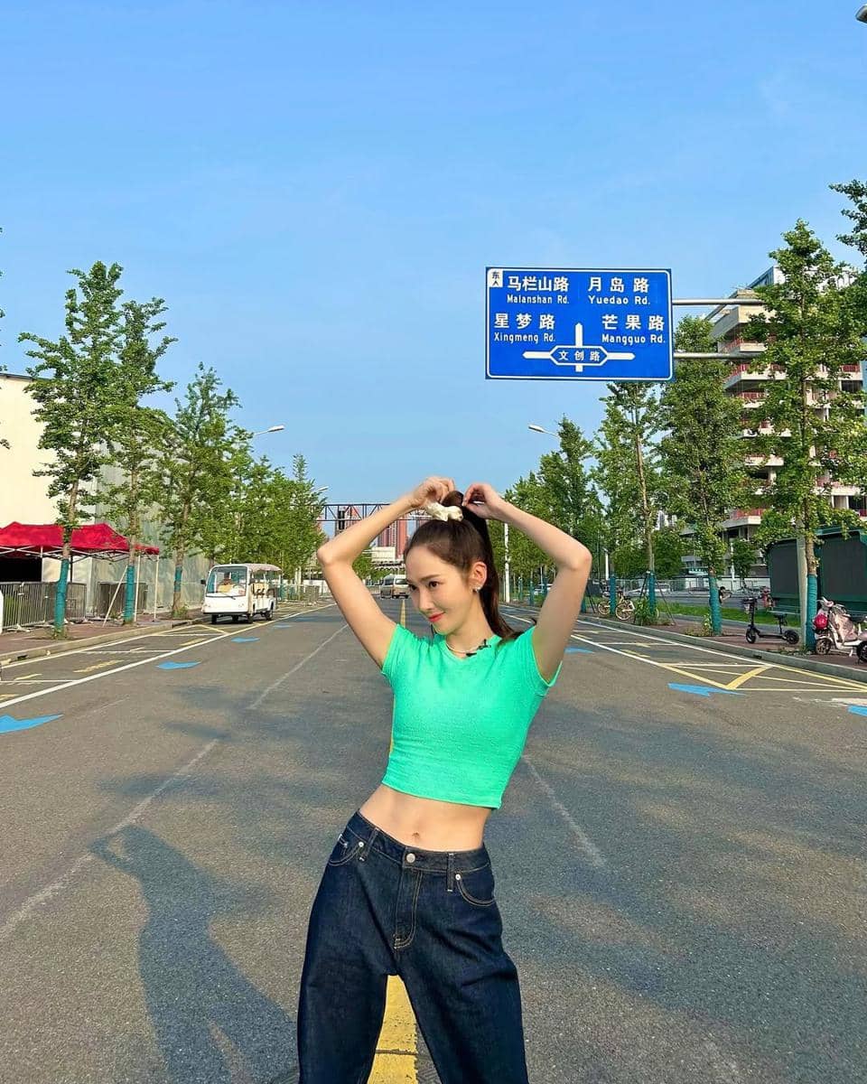 Seorang wanita mengenakan crop top hijau dan celana jeans longgar berpose di tengah jalan dengan latar pepohonan dan papan penunjuk arah.