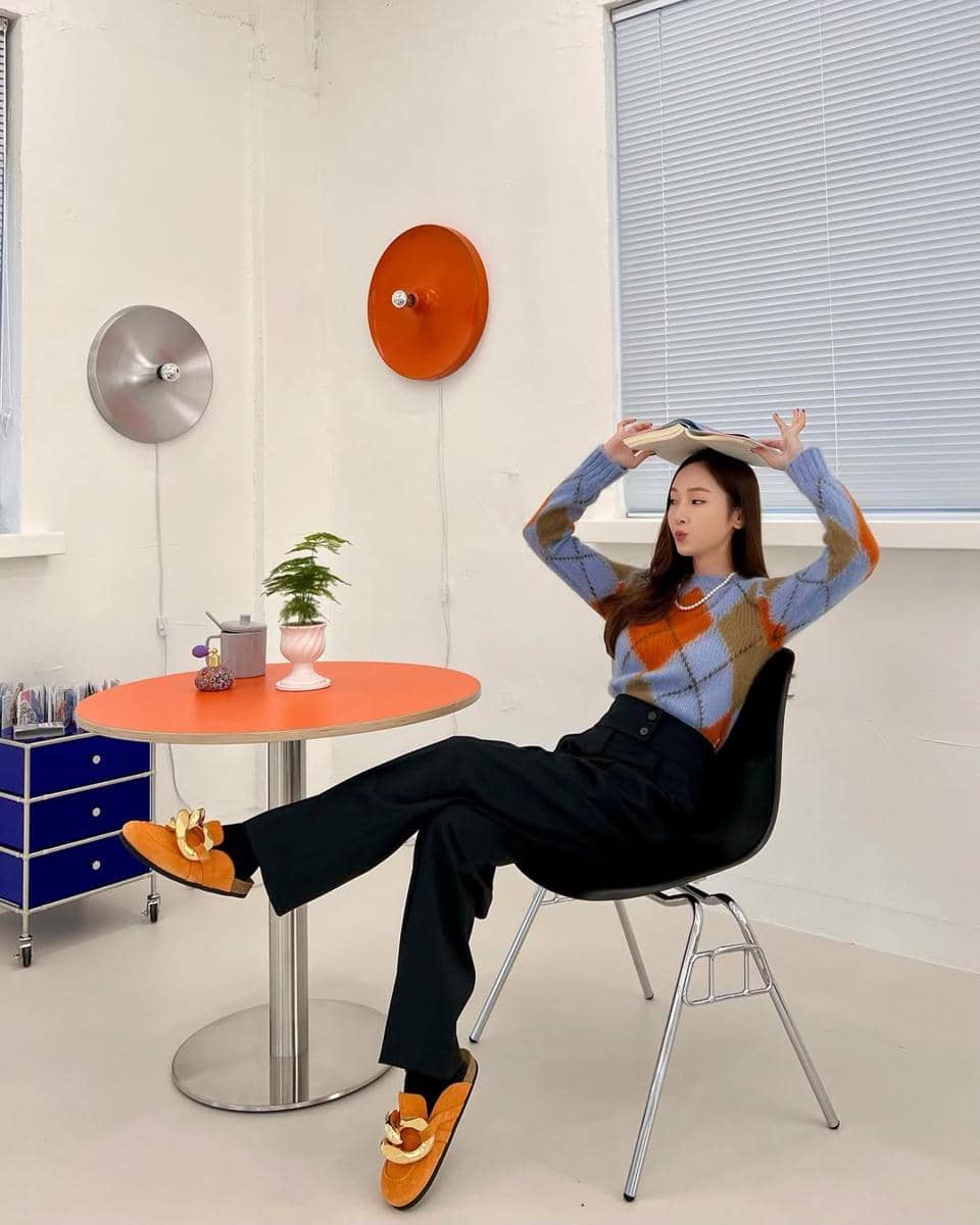 Seorang wanita duduk santai di kursi dengan kaki terangkat, mengenakan sweater bermotif kotak warna biru dan oranye serta sepatu oranye.