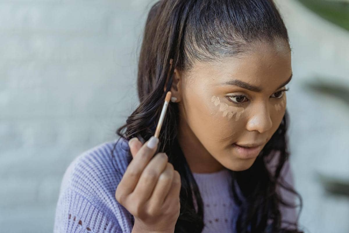 Seorang wanita sedang mengaplikasikan concealer di bawah matanya menggunakan kuas kecil untuk meratakan warna kulit wajah.