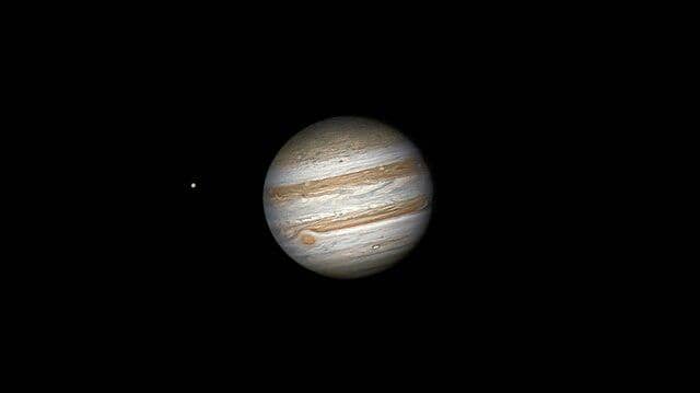 Planet Jupiter dengan pita awan berwarna oranye dan putih terlihat jelas di ruang angkasa dengan salah satu bulannya di sisi kiri.