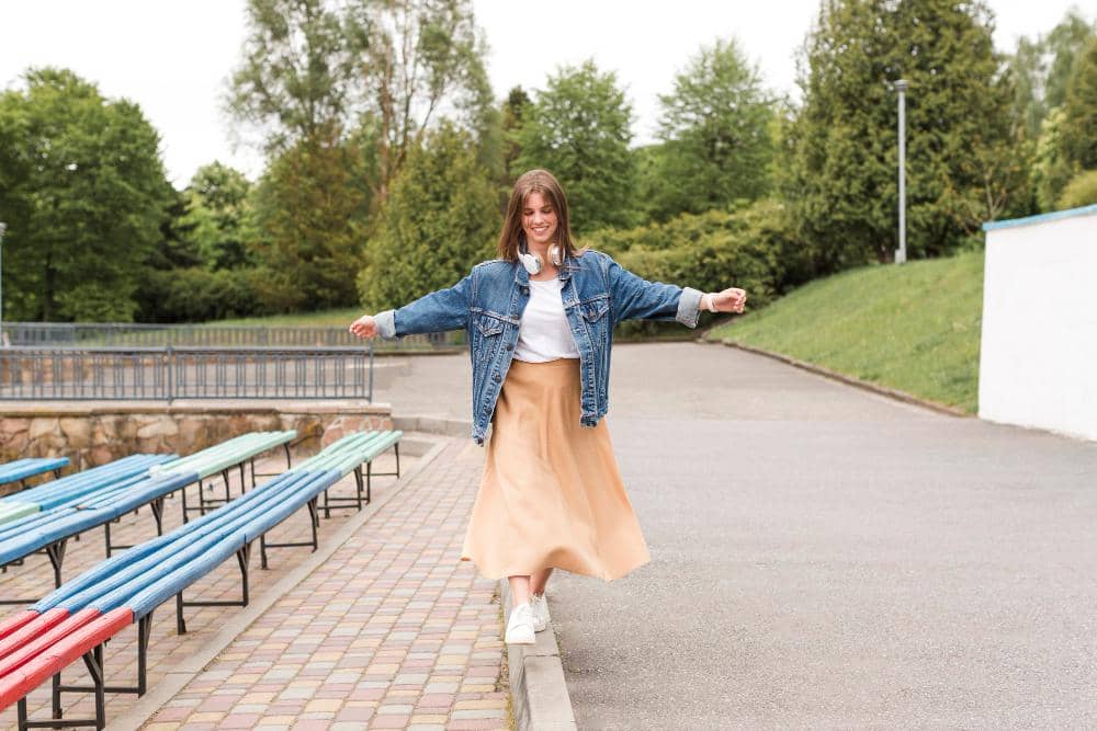 Perempuan mengenakan jaket denim dan rok panjang berjalan santai di tepi trotoar dengan latar taman hijau dan bangku warna-warni.