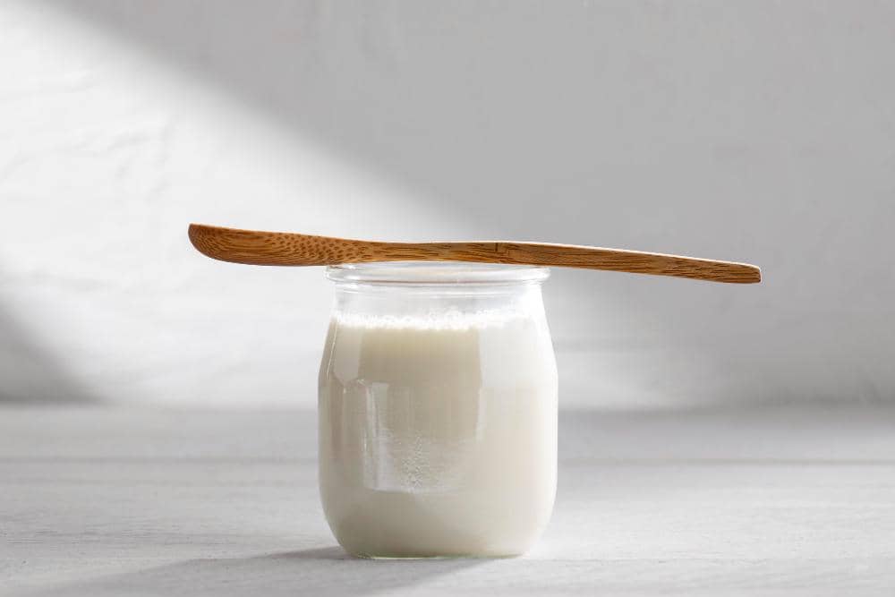 Sebuah toples kaca berisi yogurt putih dengan sendok kayu diletakkan di atasnya di atas permukaan meja berwarna abu-abu terang.