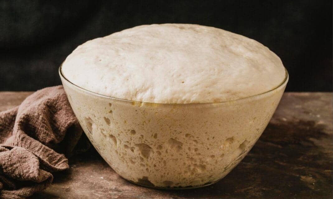 Adonan roti yang mengembang sempurna di dalam mangkuk kaca besar di atas meja dapur dengan kain cokelat di sampingnya.