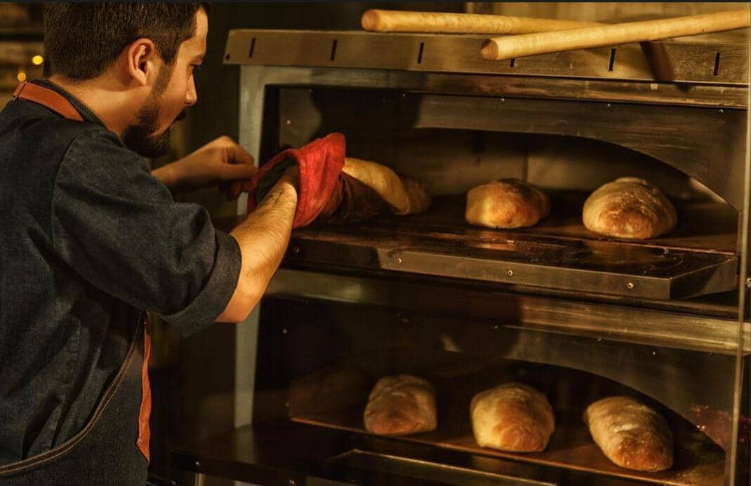 Seorang pembuat roti mengenakan sarung tangan oven sedang mengeluarkan roti panggang dari oven industri di dapur hangat.