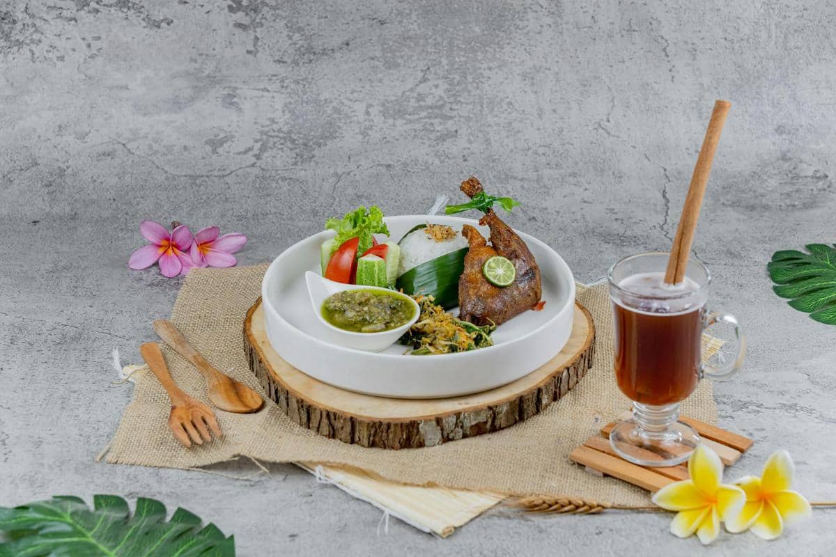 Satu piring berisi nasi, ayam goreng, sambal hijau, sayuran, dan lalapan disajikan dengan minuman teh hangat di meja dekoratif.