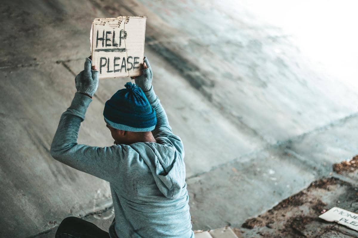 Seseorang duduk di bawah jembatan sambil mengangkat kardus bertuliskan “HELP PLEASE”, mengenakan jaket, topi rajut, dan sarung tangan, menggambarkan kondisi kerentanan, kemiskinan, dan permohonan bantuan.