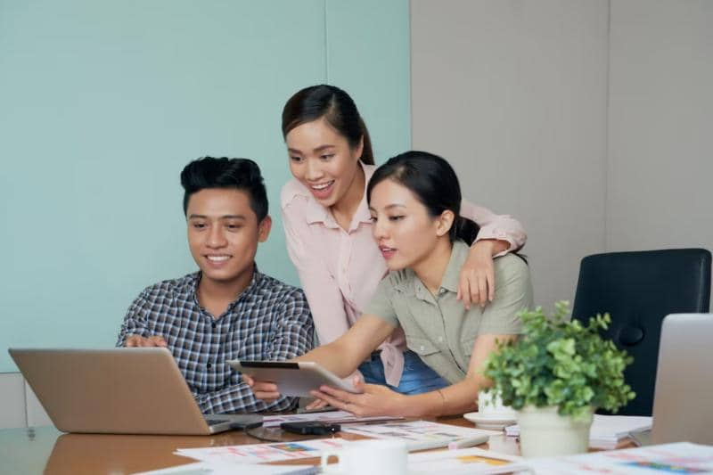 Tiga anak muda Gen Z berdiskusi dan mengelola bisnis menggunakan laptop dan tablet di ruang kerja dengan suasana kolaboratif.