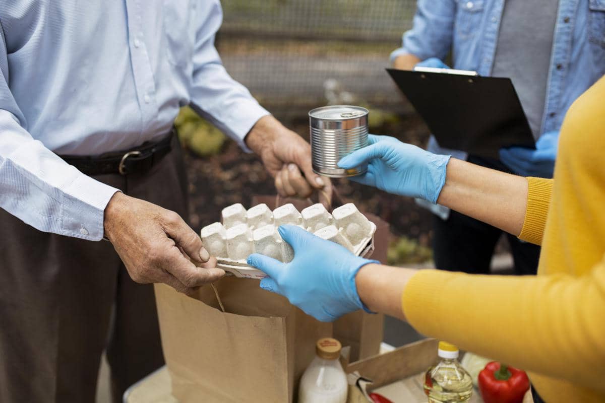 Relawan mengenakan sarung tangan menyerahkan kaleng makanan dan kemasan telur kepada seorang warga. Di atas meja terlihat tas kertas berisi bahan pokok seperti susu, minyak goreng, dan sayuran, menggambarkan kegiatan bantuan pangan dan solidaritas komunitas.