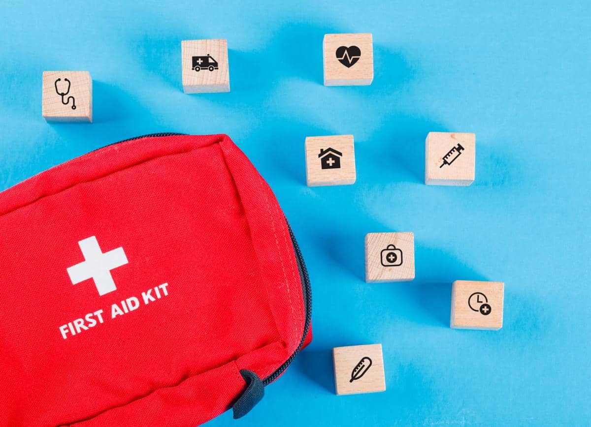 Tas P3K berwarna merah dengan tanda silang putih dan tulisan “First Aid Kit”, dikelilingi balok kayu kecil bergambar ikon layanan kesehatan seperti stetoskop, ambulans, jantung, rumah sakit, suntikan, obat, dan jam, di atas latar biru.