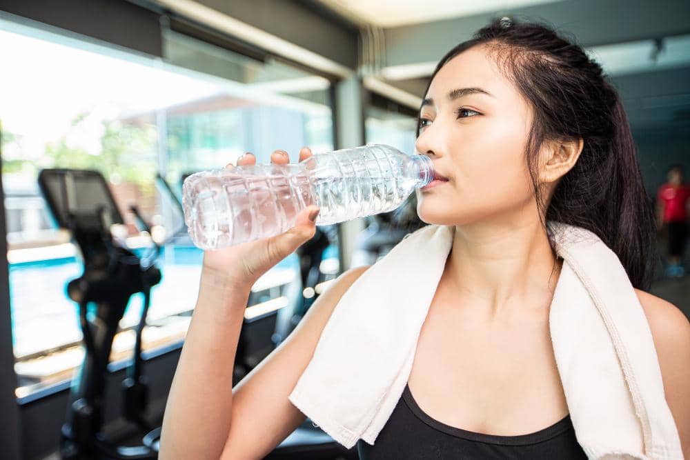 Perempuan mengenakan handuk di leher sedang minum air putih dari botol plastik di area gym dengan peralatan olahraga di belakangnya.