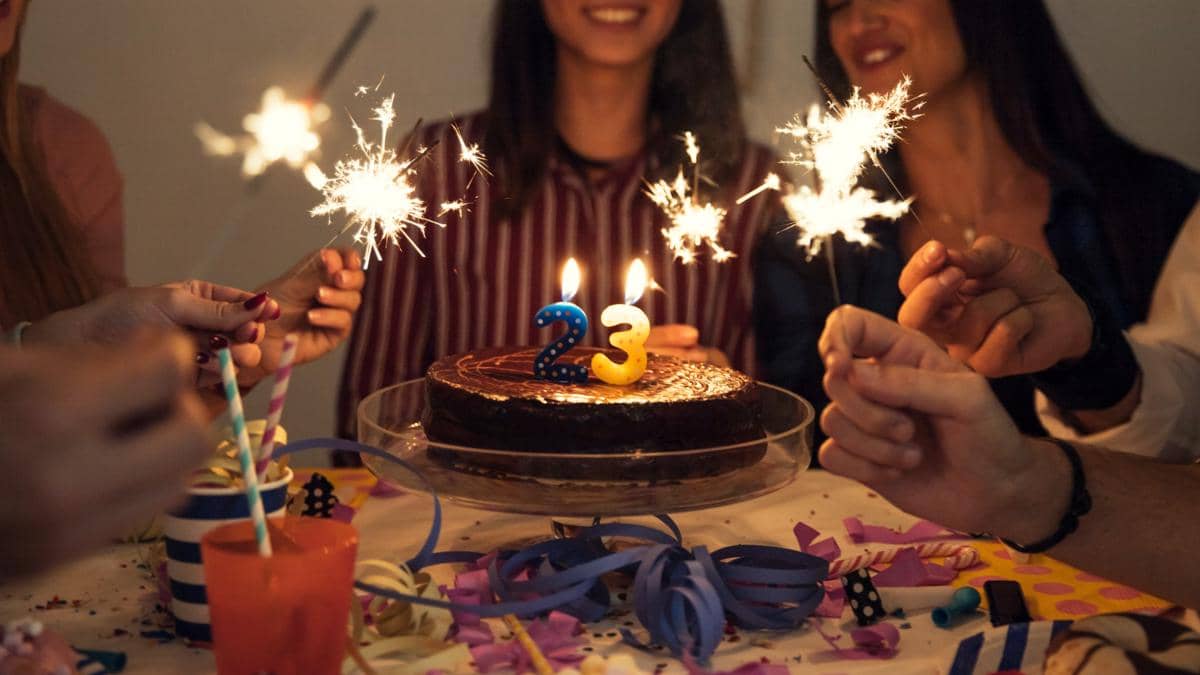 Sekelompok orang merayakan ulang tahun dengan kue cokelat bertuliskan angka 23. Lilin menyala di atas kue, sementara beberapa tangan memegang kembang api kecil. Suasana pesta tampak hangat, meriah, dan penuh kebersamaan.