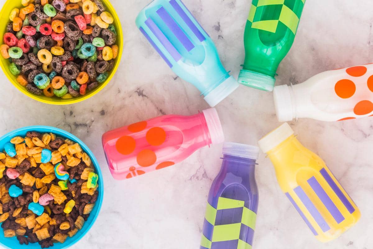 Mangkuk berisi sereal warna-warni dan botol minuman berwarna cerah dengan pola geometris di atas meja marmer.