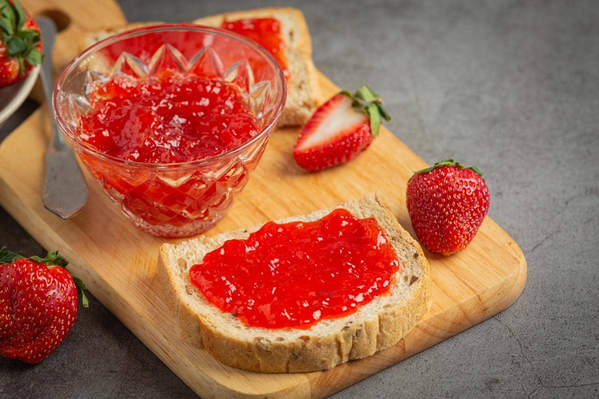 Ilustrasi selai stroberi berwarna merah dalam toples kaca, disajikan bersama stroberi segar dan roti.