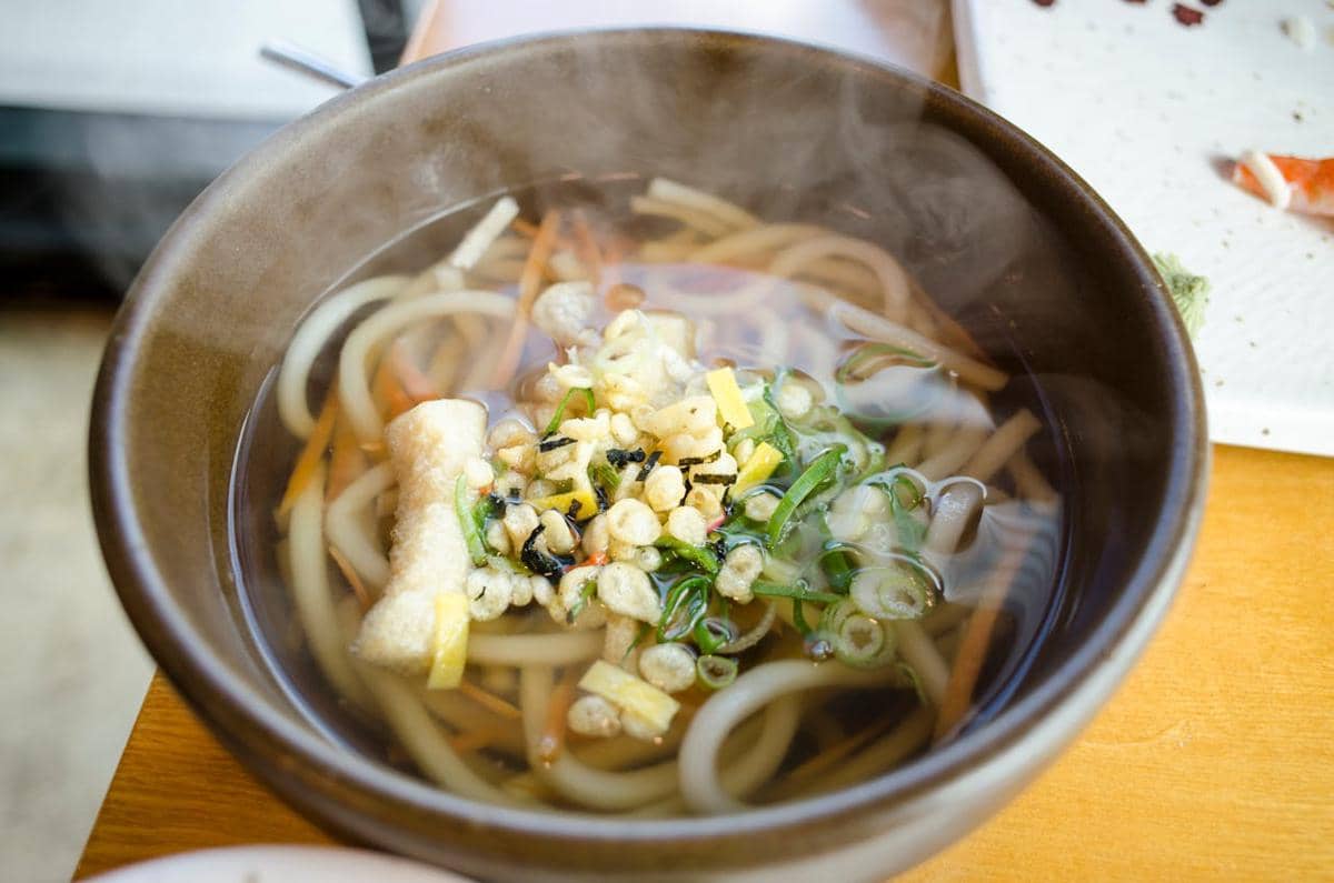 Semangkuk niku udon hangat dengan kuah bening, potongan daun bawang, tempura crumbs, dan irisan sayuran di atas meja kayu.