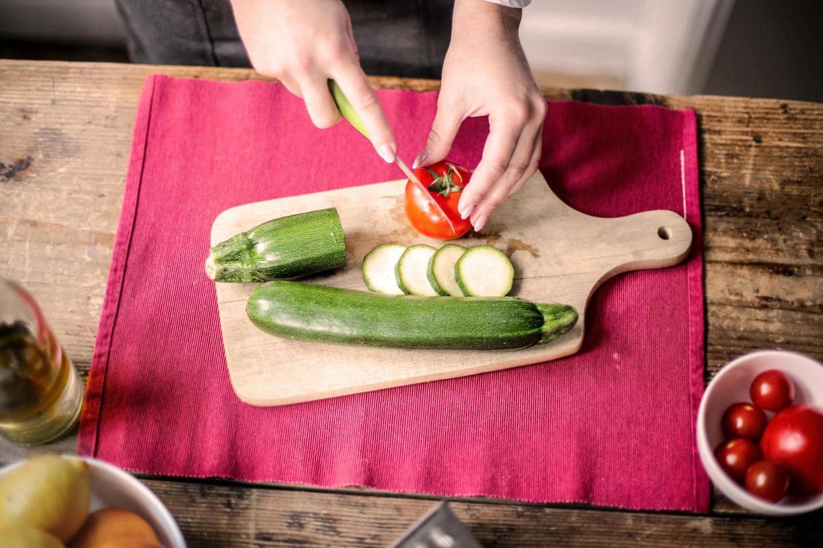 Seseorang memotong tomat di atas talenan kayu bersama potongan zucchini di dapur dengan alas kain merah.
