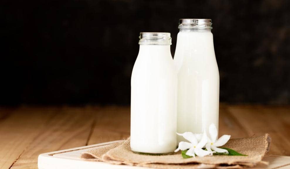Dua botol kaca berisi susu putih diletakkan di atas kain goni di meja kayu dengan latar belakang gelap dan bunga putih di depannya.