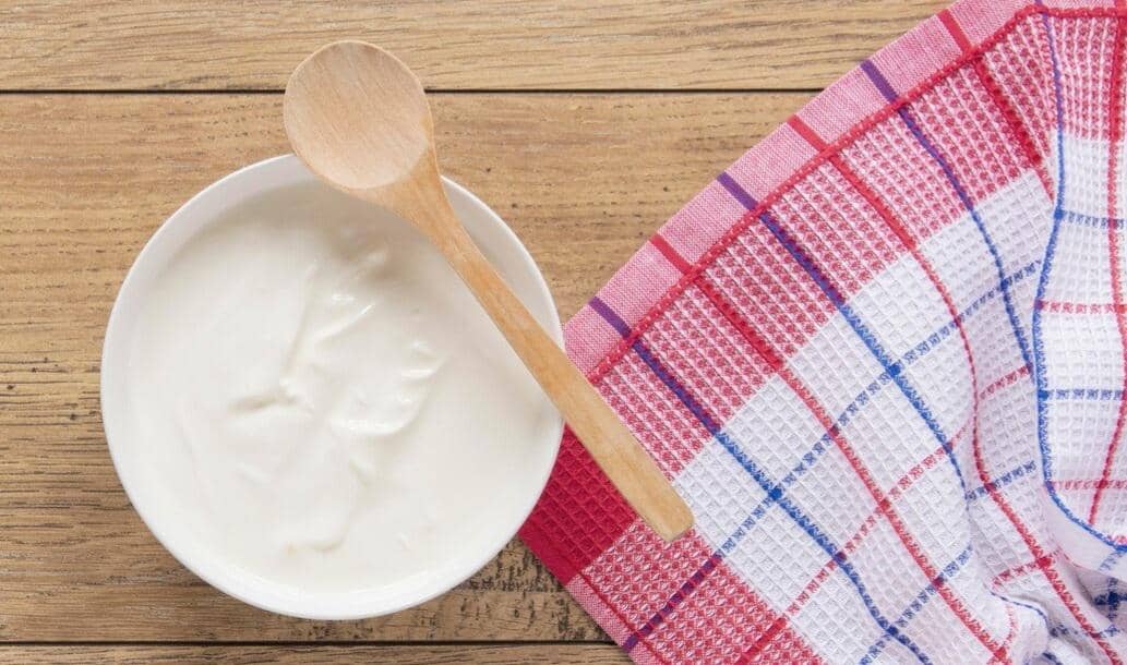 Semangkuk yogurt putih dengan sendok kayu di atas meja kayu, di samping kain lap bermotif kotak merah biru putih.
