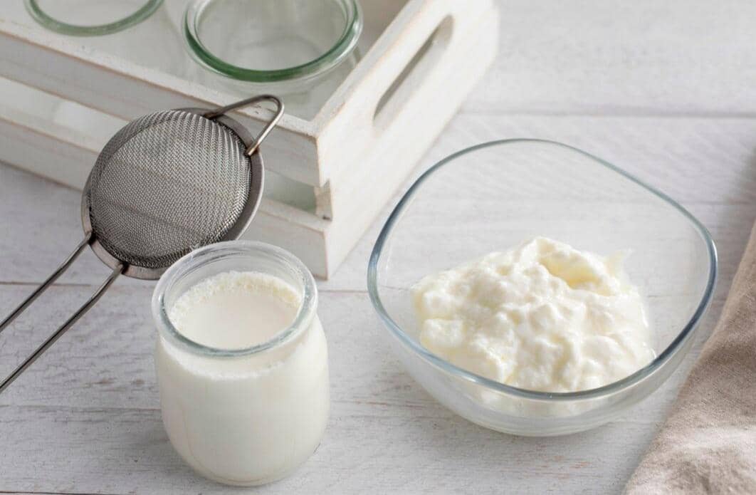 Yogurt putih dalam mangkuk kaca dan toples kecil di atas meja kayu dengan saringan logam dan wadah kaca di latar belakang.