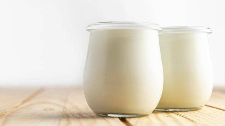 Dua toples kaca berisi yogurt putih diletakkan di atas permukaan kayu dengan latar belakang terang dan bersih.