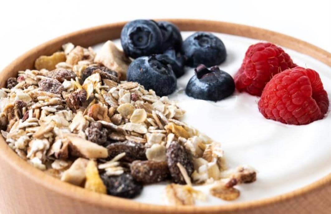 Semangkuk yogurt dengan topping granola, blueberry, dan raspberry segar tersaji dalam mangkuk kayu di atas permukaan putih.