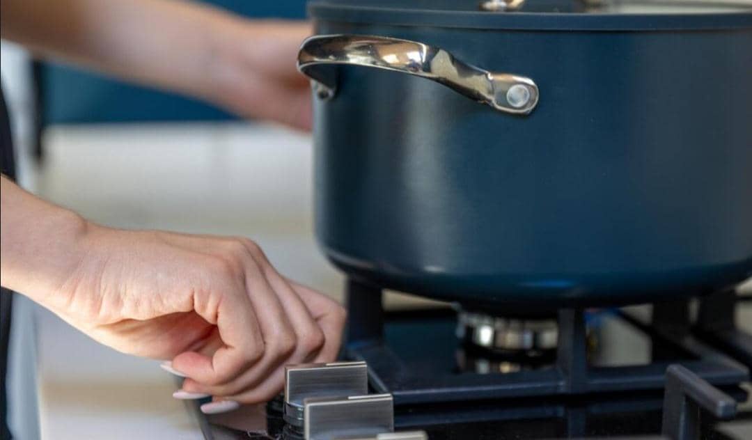Tangan seseorang sedang memutar kenop kompor gas untuk mengatur suhu saat memasak menggunakan panci berwarna biru tua.