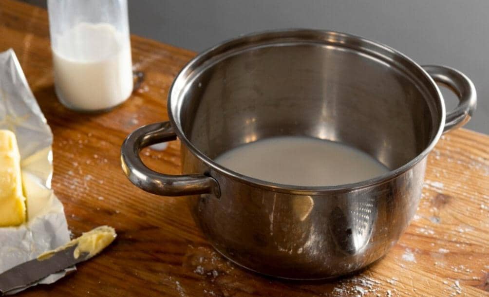 Panci stainless steel berisi cairan susu di atas meja kayu dengan sebotol susu dan mentega di sekitarnya.