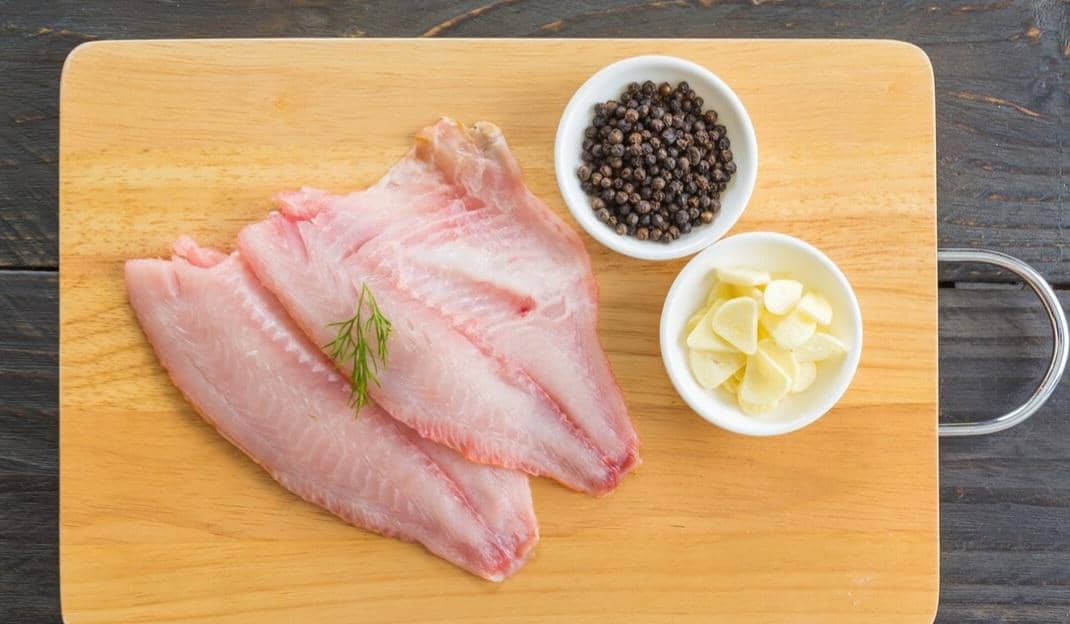 Dua potong fillet ikan mentah di atas talenan kayu dengan mangkuk berisi lada hitam dan irisan bawang putih di sampingnya.